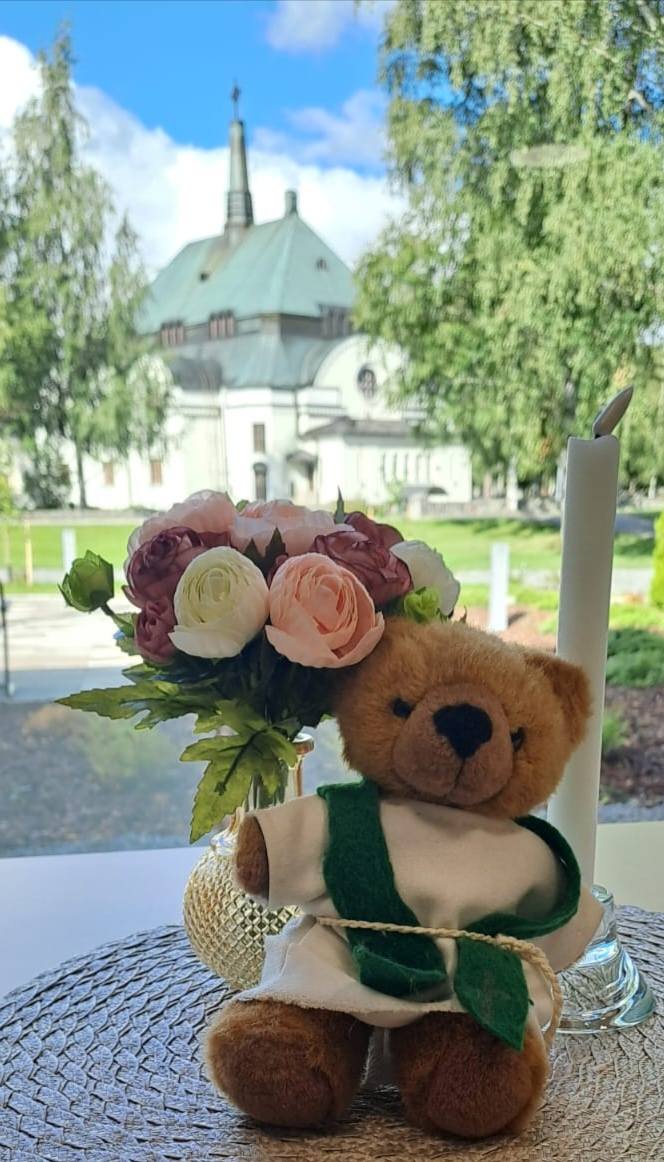 Nalle istuu pöydällä kynttilän ja kukkakimpun vieressä. Taustalla ikkuna, josta näkyy kirkko.